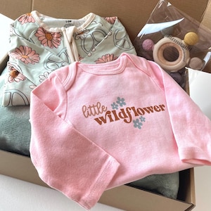 May include: A baby gift set in a cardboard box. The set includes a pink onesie with "little wildflower" text, a floral print zip-up, and a wooden teether. The onesie is long-sleeved and the zip-up has a daisy pattern.