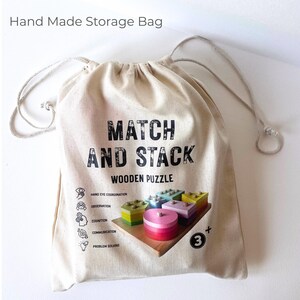 Op de afbeelding: Een beige opbergtas met trekkoord voorzien van de zwarte tekst "Hand Made Storage Bag" bovenaan en "MATCH AND STACK WOODEN PUZZLE" in het midden. De tas toont een afbeelding van kleurrijke houten vormen op een basis en iconen voor ontwikkelingsvaardigheden.
