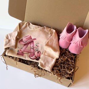 May include: A baby gift set in a cardboard box. The set includes a tan onesie with a pink cowboy hat and boot design, and a pair of pink fringed baby moccasins. The box is filled with brown crinkle paper.