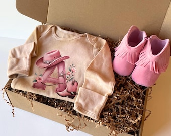 May include: A baby gift set in a cardboard box. The set includes a beige onesie with a pink cowboy hat and boot design, and a pair of pink fringed baby moccasins. The box is filled with brown shredded paper.