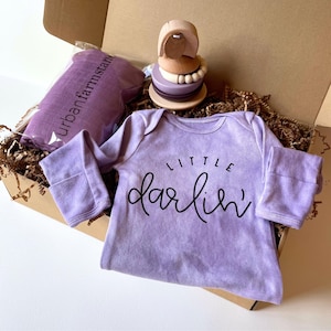Puede incluir: Un juego de regalo para bebé en una caja de cartón, que incluye un body lavanda con "Little Darlin'" en escritura negra, un juguete de madera y una manta envuelta. La caja está llena de papel de seda marrón.