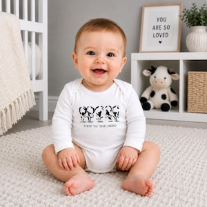 May include: A white long-sleeve baby bodysuit featuring a black graphic print of four stylized cow silhouettes in a row. Below the cows, the text reads "NEW TO THE HERD" in a simple, clean, black sans-serif font centered on the chest area.