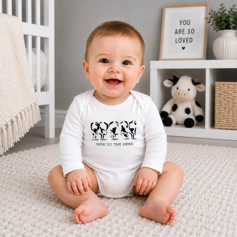 May include: A white long-sleeve baby bodysuit featuring a black graphic print of four stylized cow silhouettes in a row. Below the cows, the text reads "NEW TO THE HERD" in a simple, clean, black sans-serif font centered on the chest area.