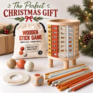 Op de afbeelding: Een houten stokjesspel met een cilindrische toren met gekleurde latten, een beige tas met de tekst "WOODEN STICK GAME", drie houten ballen, een dobbelsteen met gekleurde stippen, een ronde basis en diverse dunne houten stokjes in kleuren.
