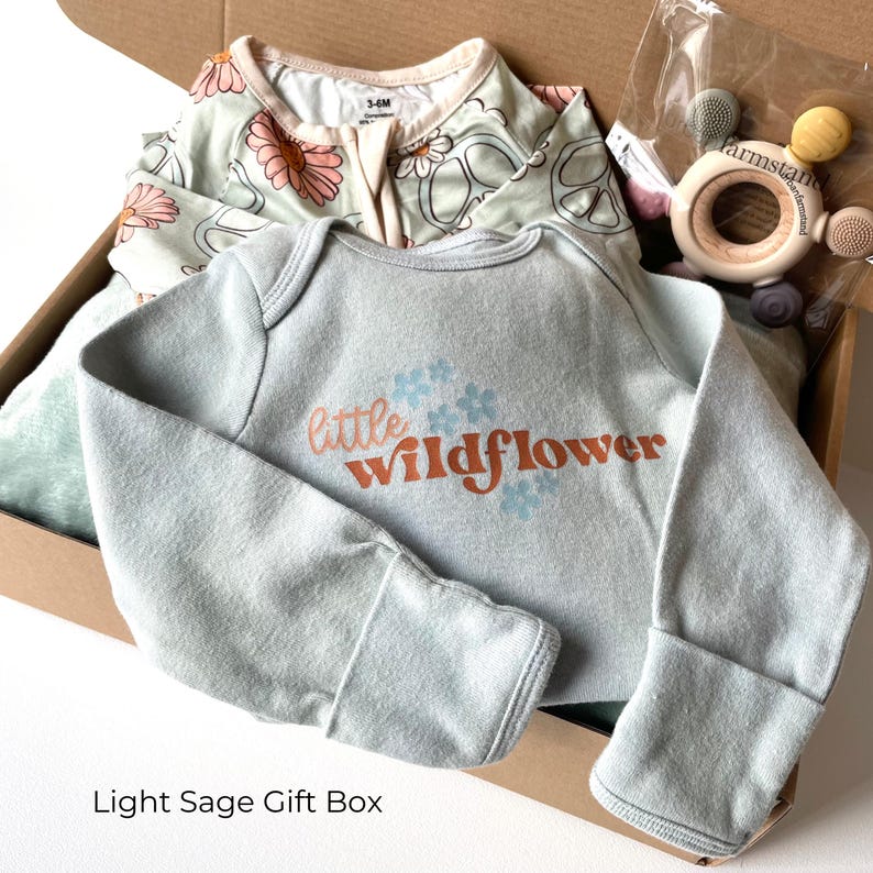 May include: A light sage gift box containing baby clothes and a wooden teether. The baby clothes include a long-sleeved bodysuit with "little wildflower" text and a floral print onesie. The teether has wooden and silicone elements.