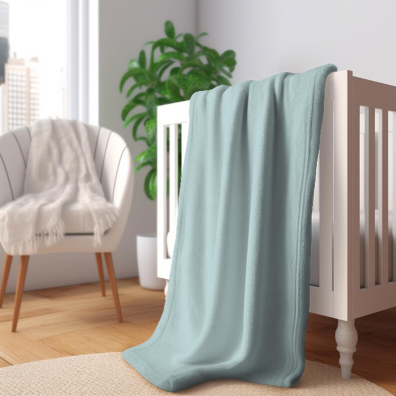 May include: A light blue fleece blanket draped over a white crib. The blanket has a soft, plush texture and a finished edge. The crib is in a bright room with a chair and a plant in the background. The blanket is a baby item.