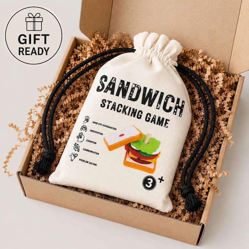 Pu&ograve; includere: Una borsa in tela color crema con coulisse che riporta il testo nero SANDWICH STACKING GAME, un'illustrazione di un panino in legno, cinque piccole icone con etichette e un'indicazione di et&agrave; 3+, all'interno di una scatola con carta di riempimento.