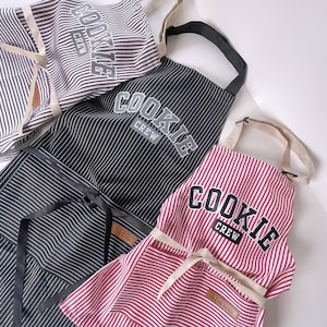 May include: Several striped aprons in black, red, and white, with the text "COOKIE CREW" printed across the chest. The aprons have adjustable neck straps and waist ties. The aprons are designed for cooking and baking.