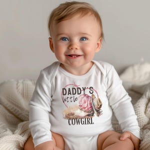 K&ouml;nnte beinhalten: Wei&szlig;er Baby-Langarmbody mit dem Text "Daddy's little Cowgirl". Die Grafik zeigt einen rosa Cowboy-Stiefel, einen braunen Cowboyhut und ein Seil. Das Baby l&auml;chelt.