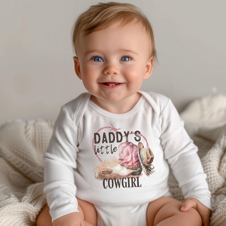 K&ouml;nnte beinhalten: Wei&szlig;er Baby-Langarmbody mit dem Text "Daddy's little Cowgirl". Die Grafik zeigt einen rosa Cowboy-Stiefel, einen braunen Cowboyhut und ein Seil. Das Baby l&auml;chelt.