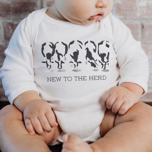 May include: A white long-sleeve baby bodysuit featuring a graphic of four stylized black cow silhouettes in a row. Below the cows, the text NEW TO THE HERD is printed in a simple black sans-serif font. The garment has a round neckline.