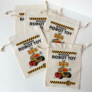 May include: Five off-white cotton drawstring bags featuring a graphic of a yellow and orange robot toy. Each bag displays the text WOODEN ROBOT TOY in black letters, accented by yellow and black hazard stripes at the top and bottom edges.