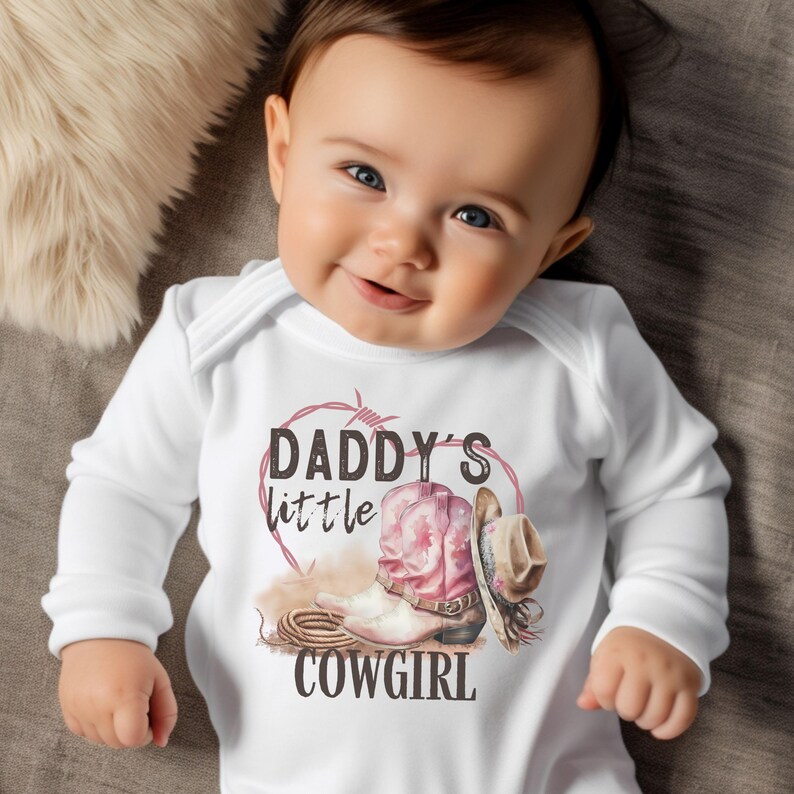 K&ouml;nnte beinhalten: Ein wei&szlig;er Baby-Body mit langen &Auml;rmeln mit einer Grafik eines rosa Cowboy-Stiefels, eines Cowboyhuts und dem Text "DADDY'S little COWGIRL" in einem herzf&ouml;rmigen Stacheldraht-Design. Das Baby l&auml;chelt.