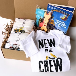 May include: A gift box with a white baby onesie printed with "NEW TO THE CREW" and a construction vehicle, a children's book, a swaddle blanket, and construction-themed toys. The box is filled with brown paper.