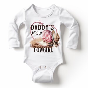 K&ouml;nnte beinhalten: Wei&szlig;er Baby-Body mit langen &Auml;rmeln. Der Body zeigt eine Grafik eines rosa Cowboy-Stiefels, eines braunen Cowboyhuts und den Text "DADDY'S little COWGIRL" in Schwarz. Ein herzf&ouml;rmiges Stacheldrahtdesign in Rot umgibt die Grafik.