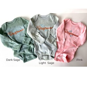 May include: Three baby onesies in dark sage, light sage, and pink. Each long-sleeved bodysuit features the text "little wildflower" in a cursive font with floral accents. The onesies have snap closures at the bottom.