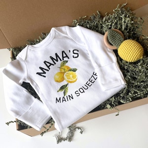 May include: White baby onesie with long sleeves, featuring the text "MAMA'S MAIN SQUEEZE" in black, along with a watercolor illustration of lemons. A yellow crocheted lemon and a wooden and green teether are also in the image, inside a cardboard box.
