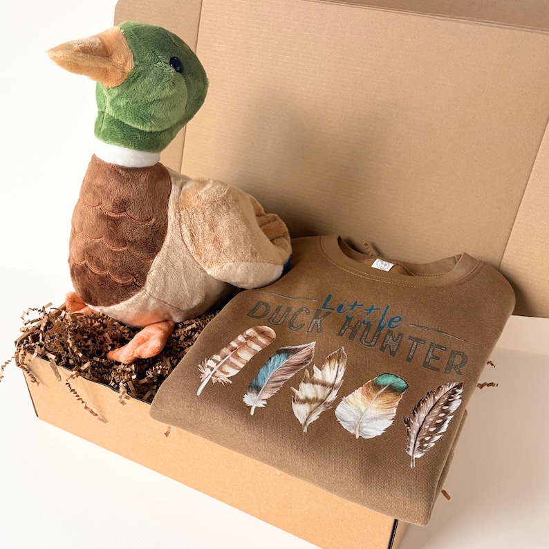 May include: A brown cardboard box contains a plush duck toy and a brown sweatshirt. The duck has a green head, brown body, and orange beak. The sweatshirt features the text "Little Duck Hunter" and feather graphics.