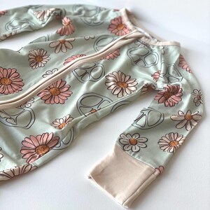 May include: A light green baby sleeper with a daisy and peace sign pattern. The sleeper has long sleeves with light pink cuffs and a full-length zipper. The daisies are pink and tan.