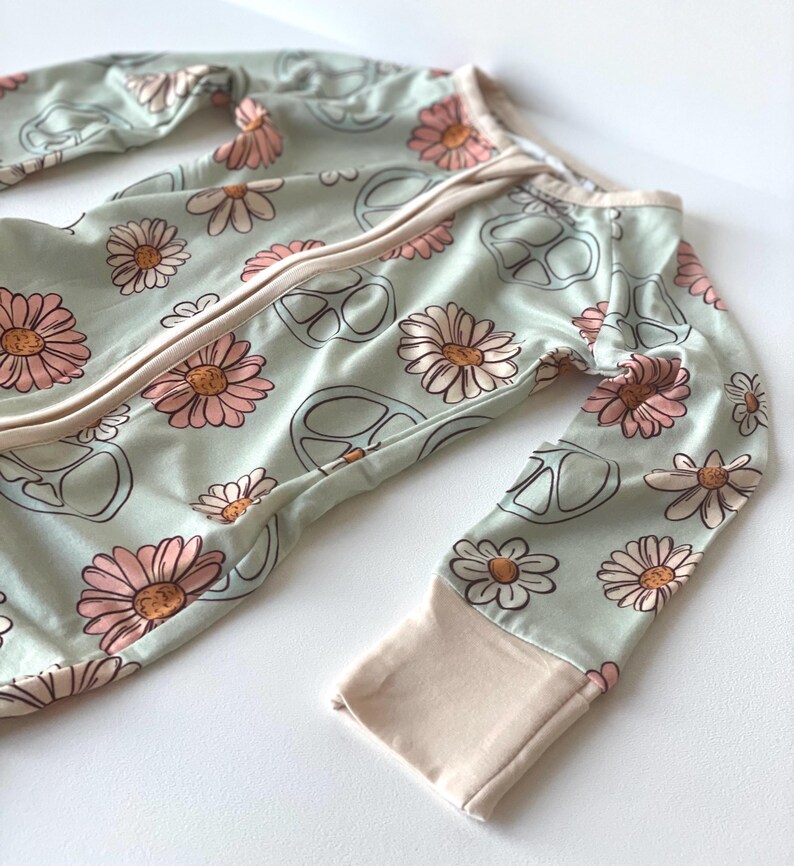 May include: A light green baby sleeper with a daisy and peace sign pattern. The sleeper has long sleeves with light pink cuffs and a full-length zipper. The daisies are pink and tan.