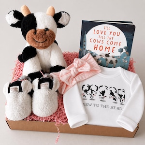 May include: A baby gift set featuring a plush cow, a children's book titled "I'll Love You Till the Cows Come Home", a pink bow headband, a white onesie with cow graphics, and white baby booties. The set is presented in a brown box.