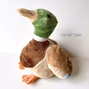 May include: A plush duck toy, approximately 10 inches tall, with a green head, brown and tan body, and orange feet. The duck has a white collar and a tan beak. Text on the image reads: "I'm 10" Tall!"