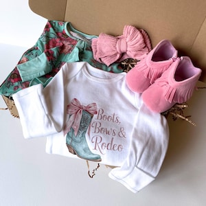 May include: A baby gift set in a cardboard box. The set includes a white onesie with a cowboy boot graphic and the text "Boots, Bows & Rodeo", a floral print top, a pink bow headband, and pink fringed baby booties.