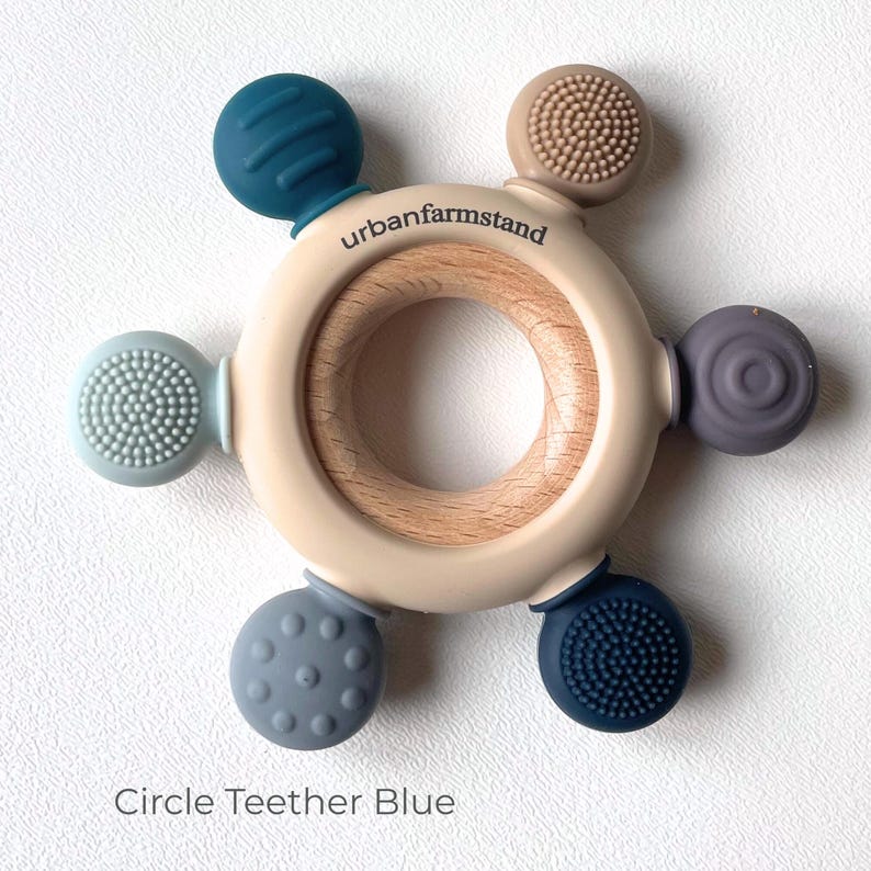 May include: A baby teether toy with a wooden center and a beige outer ring. Six colorful, textured silicone spheres in shades of blue, gray, and tan are attached around the ring. The text "urbanfarmstand" is printed on the ring, and the text "Circle Teether Blue" is at the bottom.