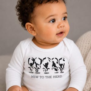 May include: A white long-sleeve baby bodysuit featuring a black graphic print of five stylized cow silhouettes standing in a row. Below the cows, the text reads NEW TO THE HERD in a simple black font. The garment has a round neckline and long sleeves.