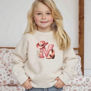 May include: A beige sweatshirt featuring a pink letter "M" with a cowboy hat and boot design. The design includes floral accents. The sweatshirt is worn by a young girl with blonde hair.