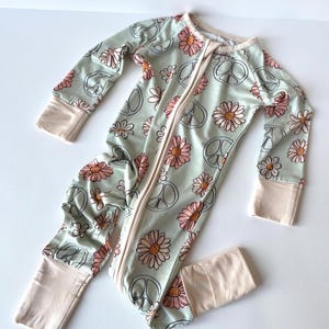 May include: A light green baby sleeper with a daisy and peace sign pattern. The long-sleeved sleeper has a full-length zipper and beige cuffs and trim. The floral design features pink and white daisies and black peace signs.