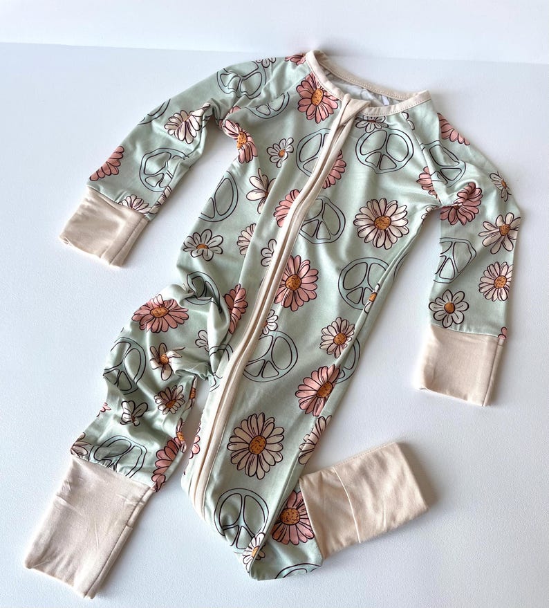 May include: A light green baby sleeper with a daisy and peace sign pattern. The long-sleeved sleeper has a full-length zipper and beige cuffs and trim. The floral design features pink and white daisies and black peace signs.
