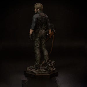 May include: A detailed figurine of a character from a video game, viewed from the back. The figure is dressed in a black shirt, dark pants, and gloves, holding a rifle. The base is a rocky terrain, and the overall color scheme is dark and realistic.