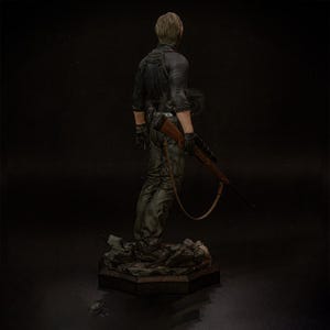 May include: A detailed figurine of a character in a black jacket and green pants, holding a rifle. The figure is standing on a textured base. The character is facing away from the viewer.