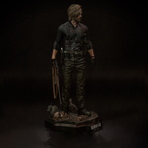 May include: A detailed figurine of a man in a black tactical outfit, holding a weapon. The figure stands on a base with the text "RESIDENT EVIL" and a skull. The figure has short, light brown hair and is looking to the side.