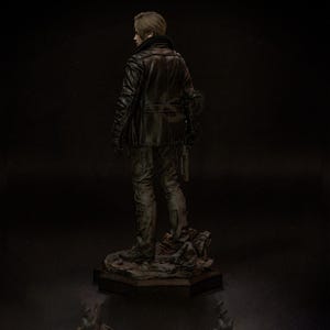 May include: A detailed statue of a character from a video game, viewed from the back. The figure wears a black leather jacket, dark pants, and gloves, holding a gun. The statue is on a rocky base, with a dark background.