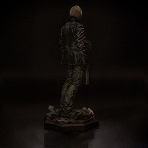 May include: A detailed figurine of a person in a black leather jacket and pants, holding a knife. The figure stands on a rocky base with a dark background. The figure is facing away from the viewer.