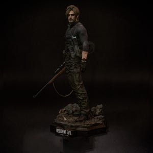 May include: A detailed figurine of a character from Resident Evil, posed with a rifle. The figure is dressed in tactical gear, with a black long-sleeve shirt and camouflage pants. The base of the figure has the text "RESIDENT EVIL" and "requiem".