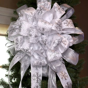 May include: A large, decorative Christmas bow made of white and silver ribbon. The bow features a layered design with silver snowflake patterns and shimmering accents. Long, flowing ribbons cascade down, adding to the festive look.