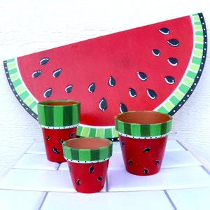 May include: Three painted terracotta pots shaped like watermelon slices. The pots are red with green stripes and black seeds. The largest pot is in the background and is partially visible. The two smaller pots are in the foreground and are fully visible.