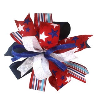 May include: A decorative bow featuring red, white, and blue ribbons. The red ribbons are adorned with white stars and blue glitter stars. White glitter and blue ribbons are also incorporated, creating a patriotic theme. The bow is suitable for various decorative purposes.