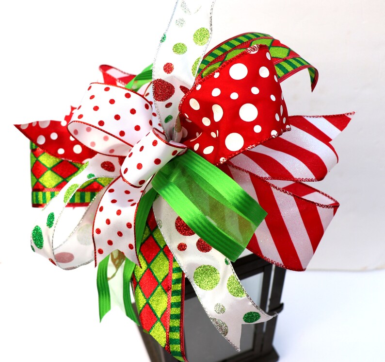Pu&ograve; includere: Un topper per lanterna di Natale festivo con un grande fiocco fatto di nastro rosso, bianco e verde con pois e strisce.