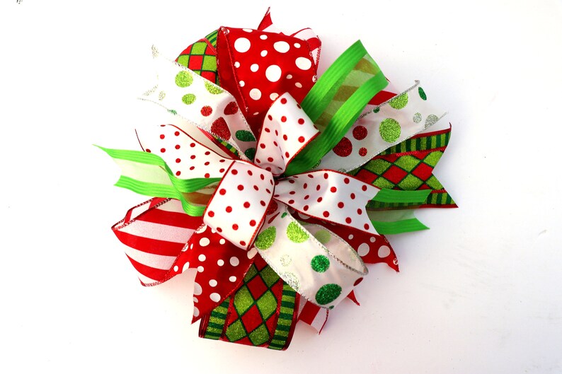 Pu&ograve; includere: Un fiocco di Natale realizzato con nastro rosso, bianco e verde. Il nastro presenta diversi motivi, tra cui pois, strisce e un motivo a quadri.