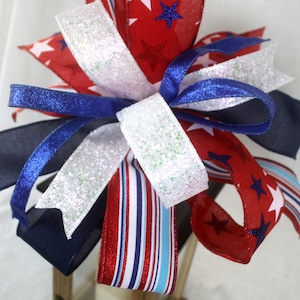 May include: A decorative bow featuring red, white, and blue ribbons. The red ribbons are patterned with white stars and blue stars. White glitter ribbons and striped ribbons add to the festive design. The bow is likely for patriotic or holiday decor.