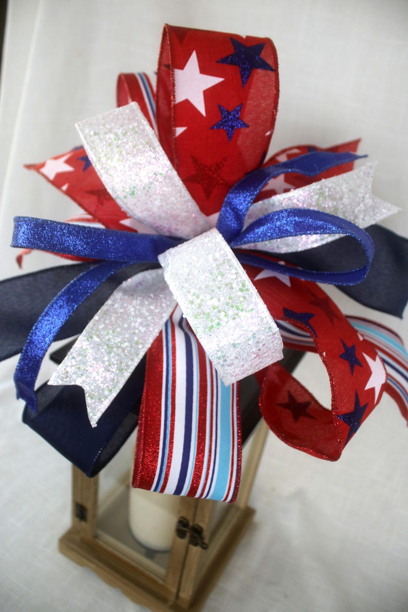 May include: A decorative bow featuring red, white, and blue ribbons. The red ribbons are patterned with white stars and blue stars. White glitter ribbons and striped ribbons add to the festive design. The bow is likely for patriotic or holiday decor.