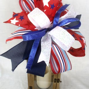 May include: A decorative bow arrangement in red, white, and blue, featuring star and stripe patterns. The bow is atop a small wooden lantern with a candle inside. The ribbons have glitter accents.