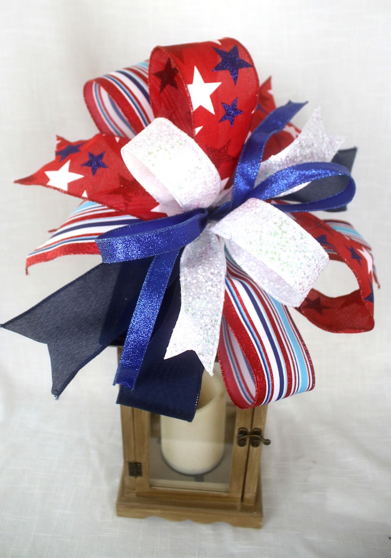 May include: A decorative bow arrangement in red, white, and blue, featuring star and stripe patterns. The bow is atop a small wooden lantern with a candle inside. The ribbons have glitter accents.
