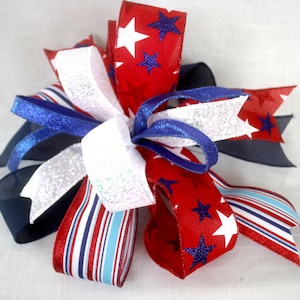 May include: A decorative bow featuring red, white, and blue ribbons. The red ribbons are adorned with white stars and blue glitter stars. Other ribbons are white with glitter, blue, and red and white stripes. This patriotic bow is suitable for various decorative uses.
