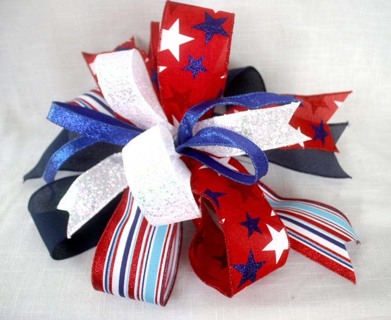 May include: A decorative bow featuring red, white, and blue ribbons. The red ribbons are adorned with white stars and blue glitter stars. Other ribbons are white with glitter, blue, and red and white stripes. This patriotic bow is suitable for various decorative uses.
