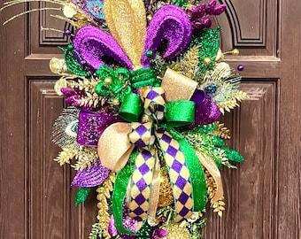 Puede incluir: Una festiva corona de puerta de Mardi Gras con acentos morados, verdes y dorados. Incluye plumas de pavo real, cintas brillantes y adornos decorativos. La corona se exhibe en una puerta de madera marrón.